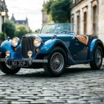 A classic British roadster parked on a cobblestone street, surrounded by historic architecture and lush greenery