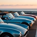 A row of classic American roadsters parked along a scenic coastal highway at sunset, with the ocean in the background and a warm, nostalgic glow